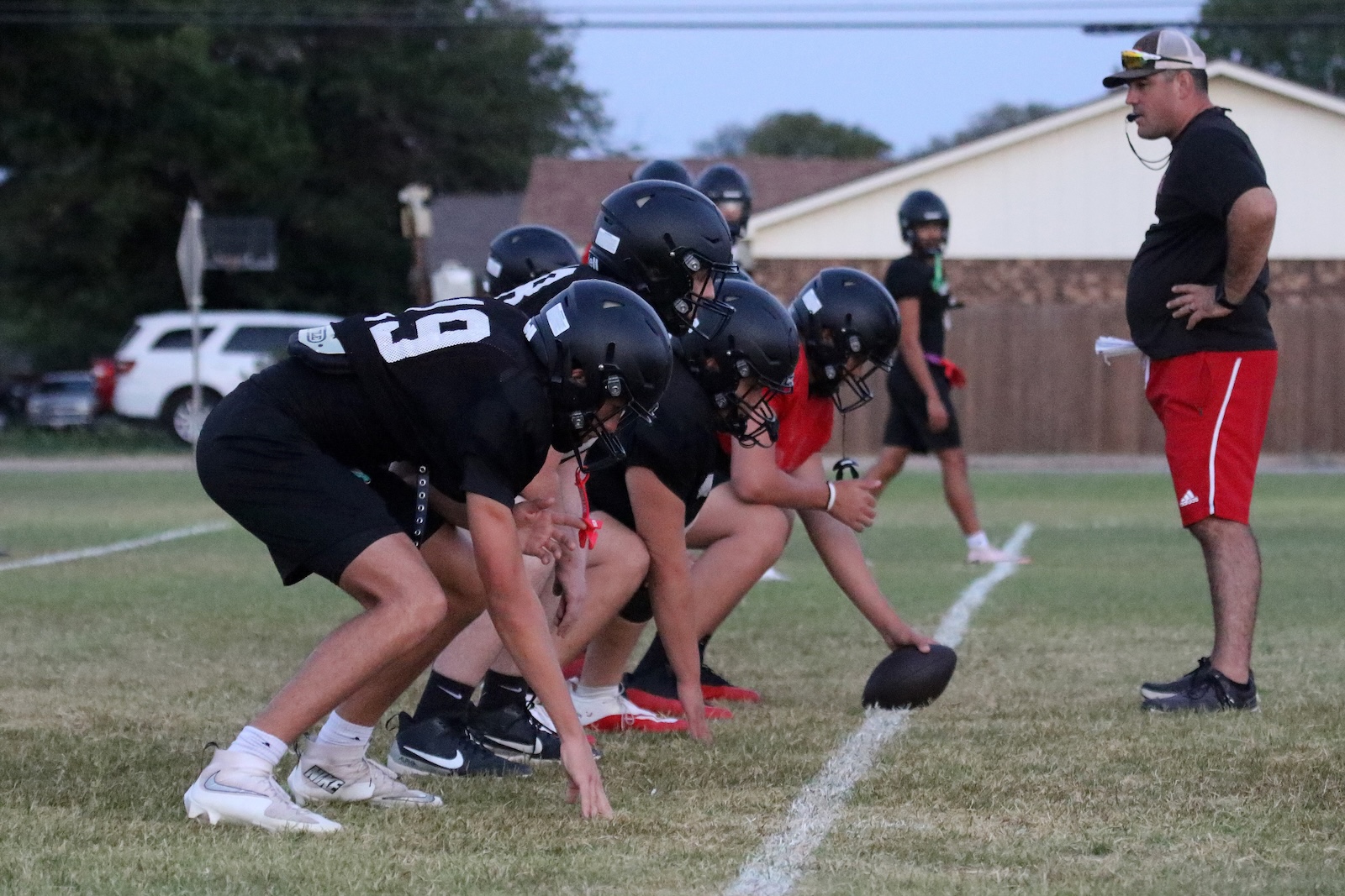Group of football players in black uniforms crouched at the line of scrimmage, coach standing to the right observing on the field.