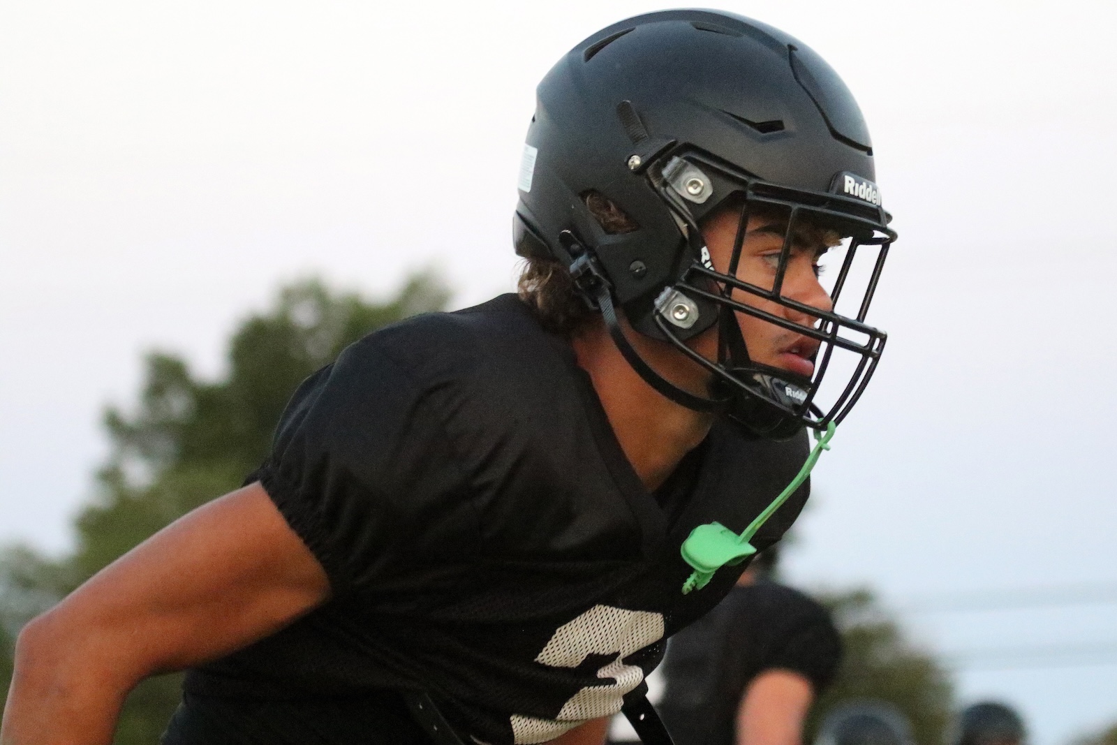 Football player in a black helmet and jersey, leaning forward during a play with a green mouthguard visible near his chin strap