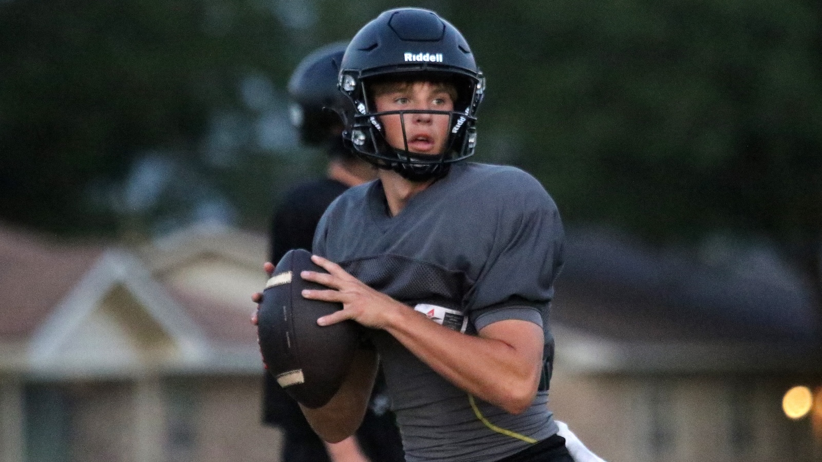 Young football quarterback in a gray jersey and black Riddell helmet grips the ball, eyes scanning for a receiver.