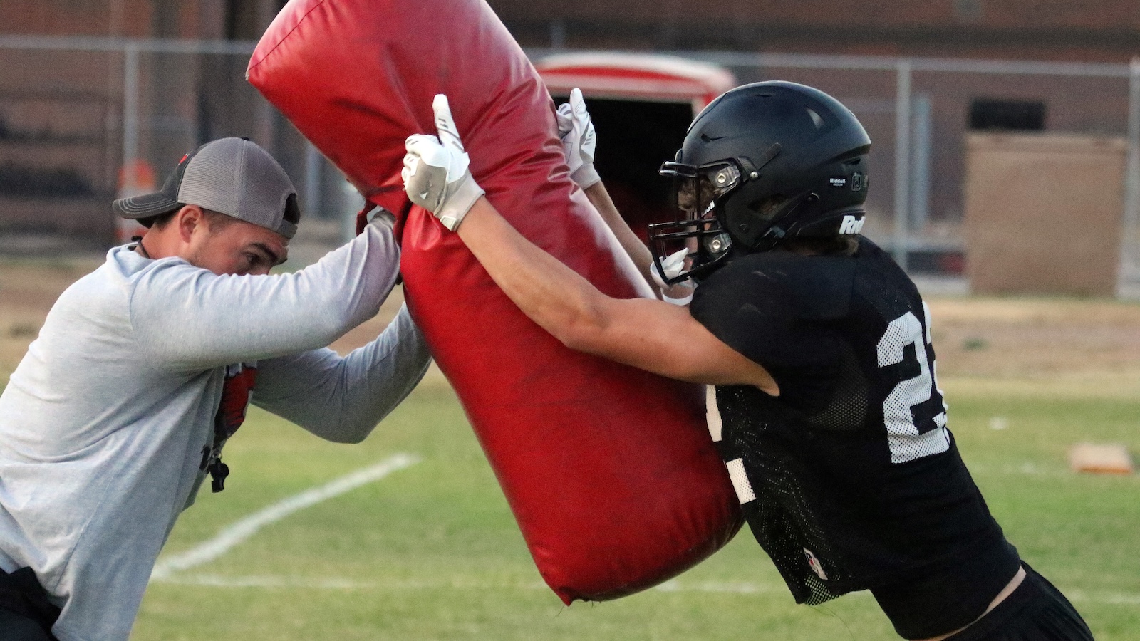 Two football players push a large red tackling dummy during practice on a grassy field.