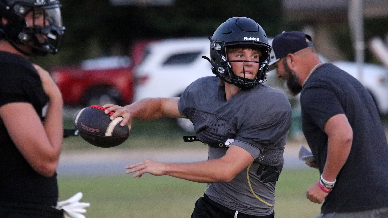 Quarterback in a gray shirt and black helmet throws a football on a grass field, with teammates nearby.