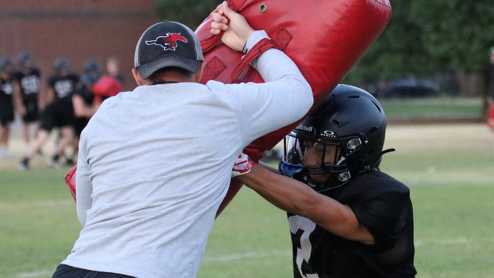 Coach in a gray shirt and cap pushes a red tackling dummy while a helmeted player blocks in football practice on a grassy field.