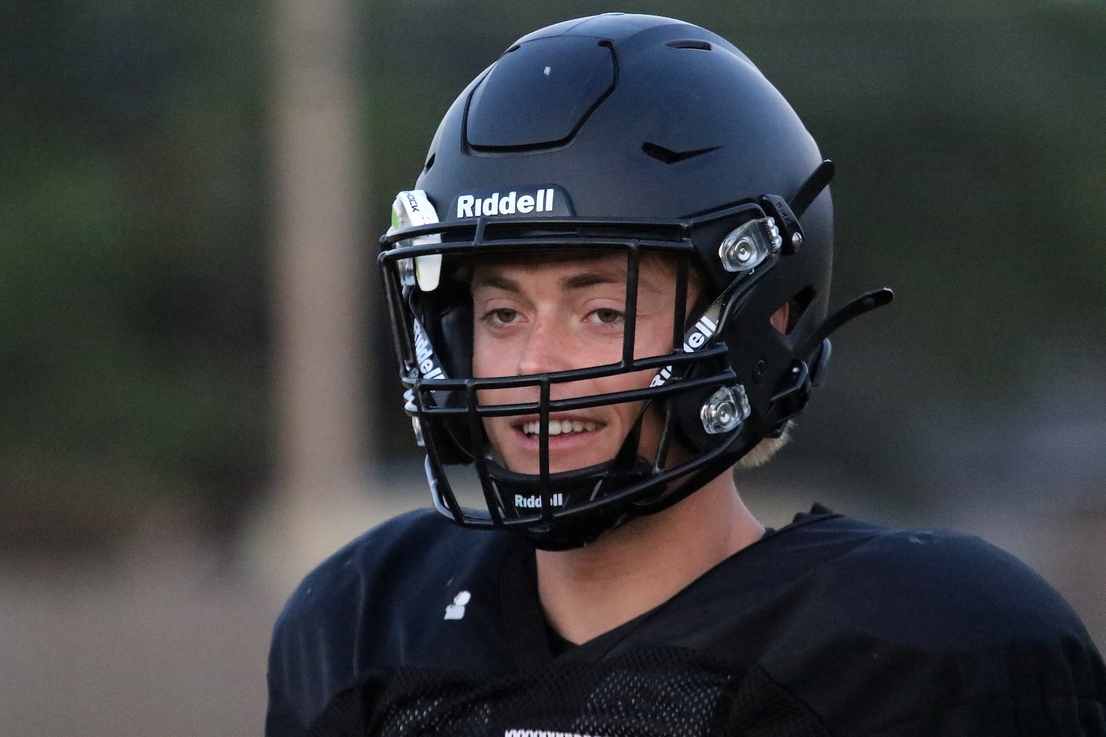Young football player wearing a black Ridell helmet and jersey, smiling at the camera on a field.