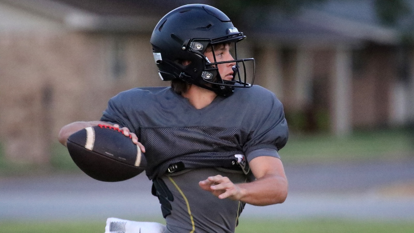 Young football player in a gray jersey and black helmet holding a football, looking to throw on a field