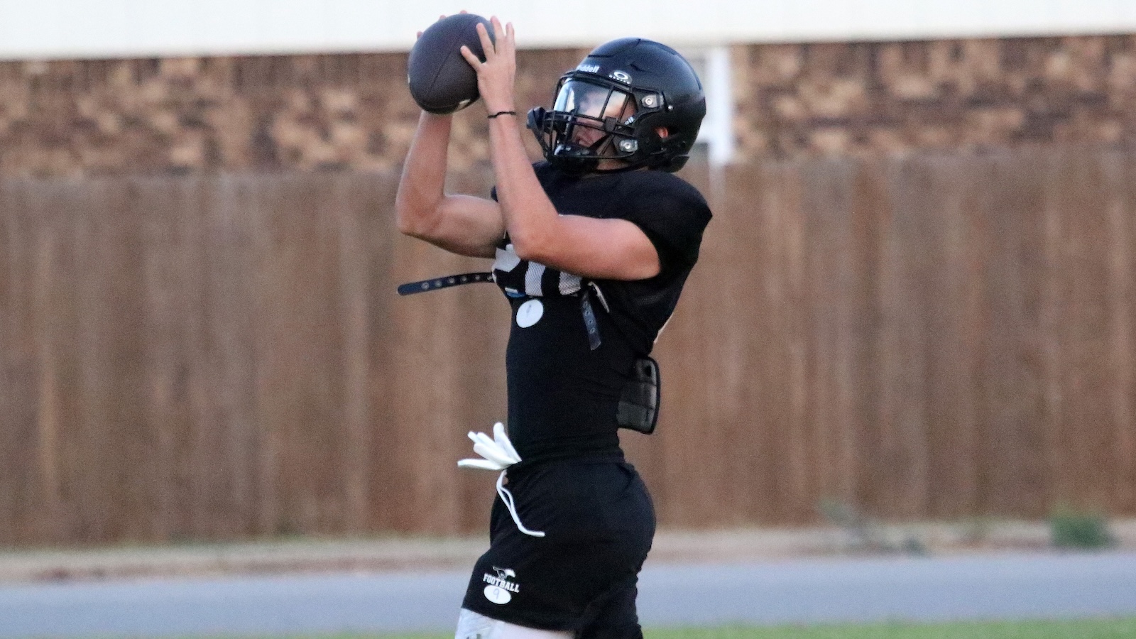 Football player in a black uniform and helmet catching a football with both hands on the field.