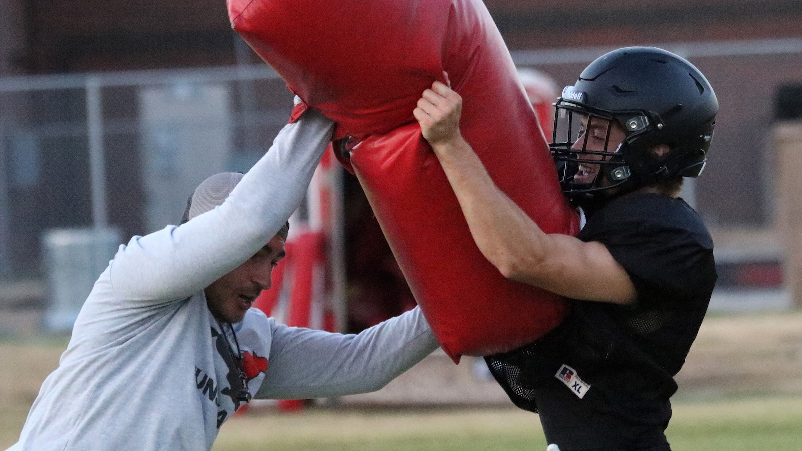Two football players train with a large red tackling dummy; one in a gray hoodie pushes it while the other in a black helmet grabs the pad.