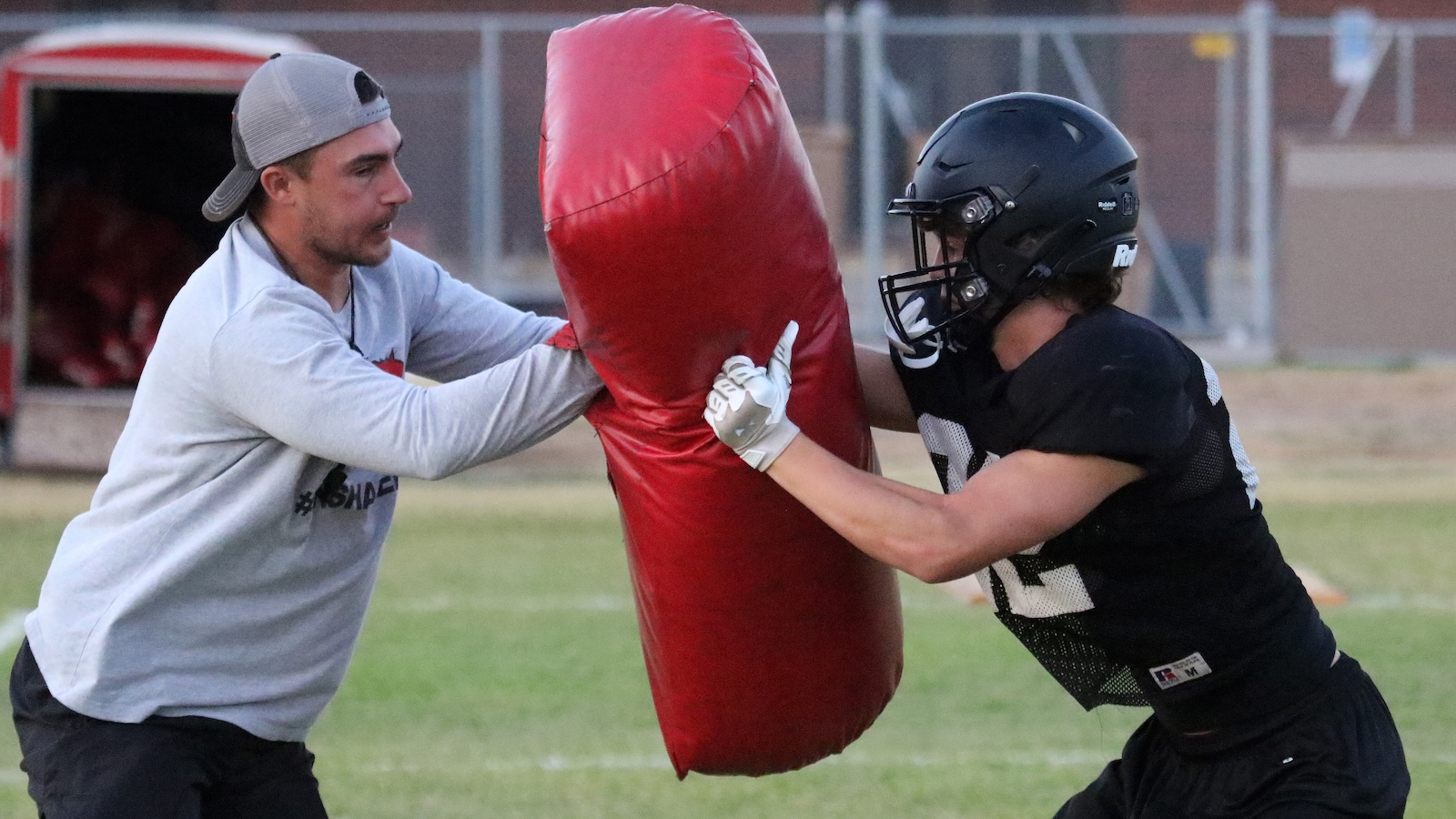 Two football players practice blocking drills on a grassy field, pushing a large red padded shield together.