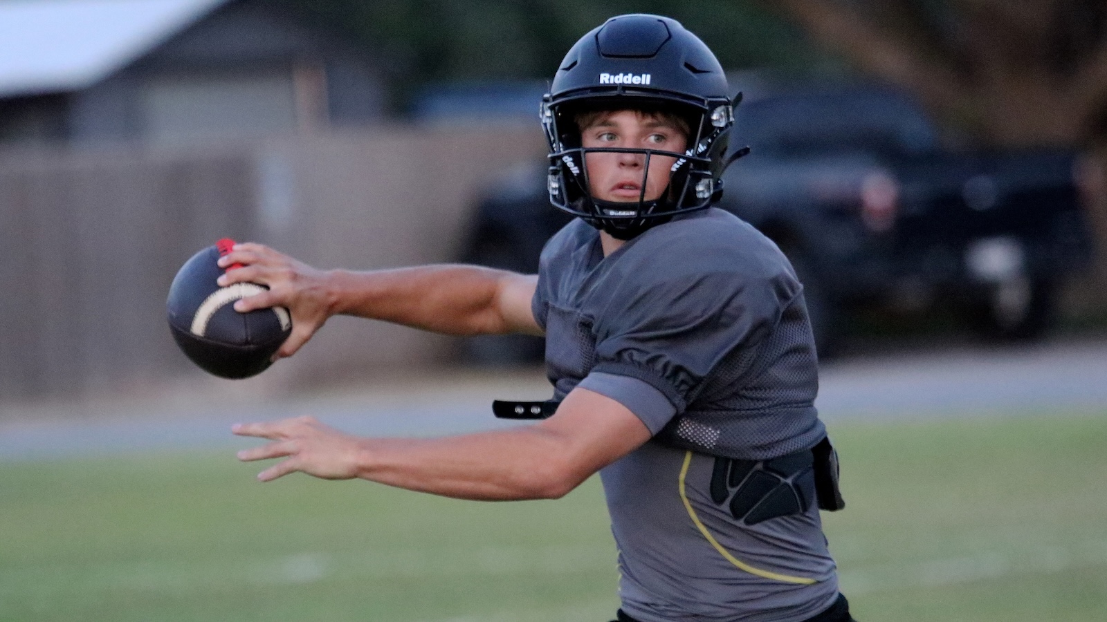Football quarterback in gray jersey throws a ball on an outdoor field, wearing a black Riddell helmet and protective gear.