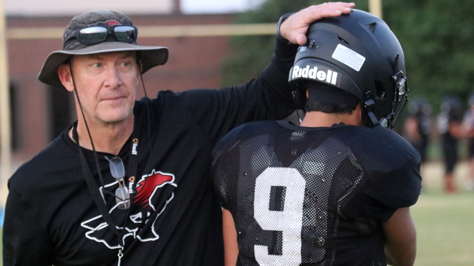 Coach resting hand on helmet of football player wearing number 9 during practice on a grassy field, coach in camo hat and sunglasses authoritatively guiding him.