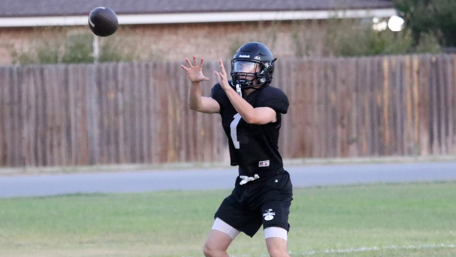 Football player in a black jersey (number 7) and helmet reaching to catch a football on a grassy field.