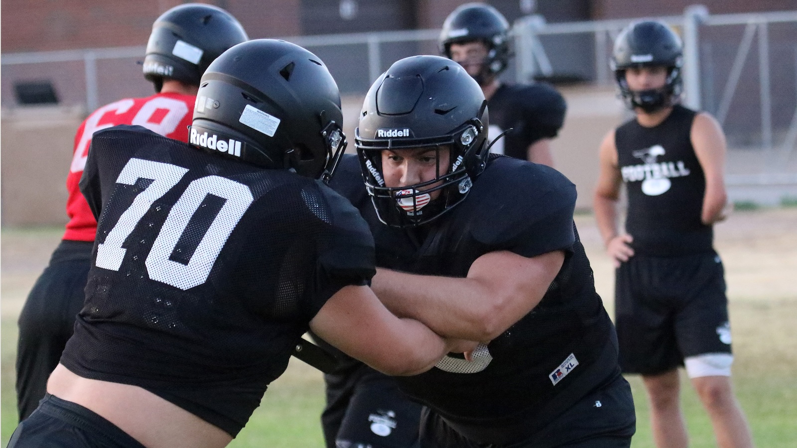 Two football players in black uniforms lock arms in a blocking drill on a practice field.