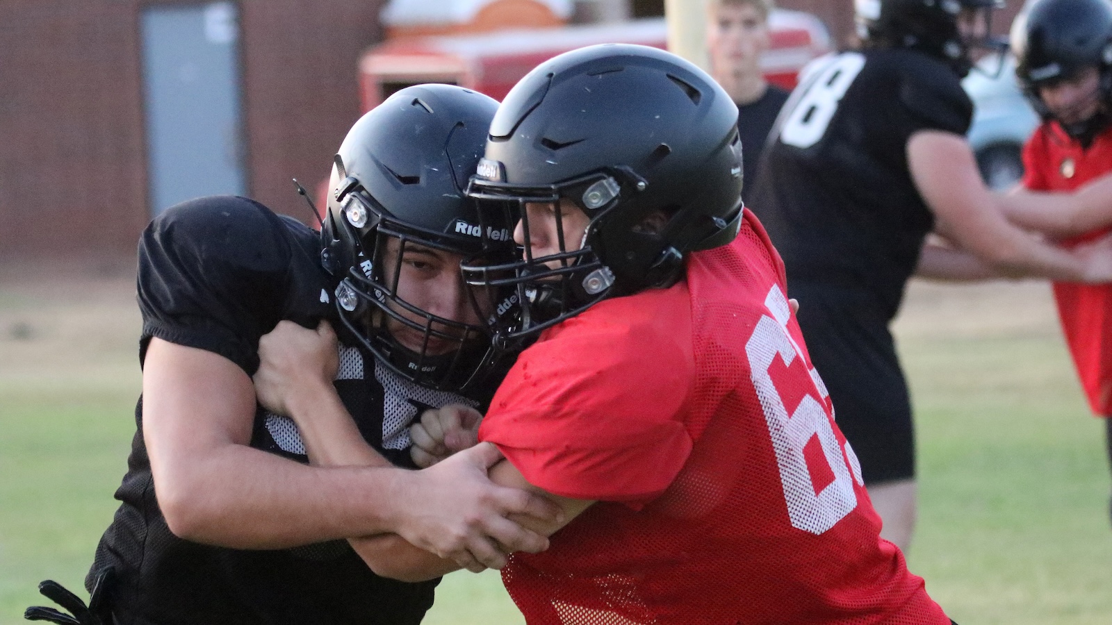 Two football players in black and red jerseys grapple each other on a grassy field, helmets clashing as they struggle for control.