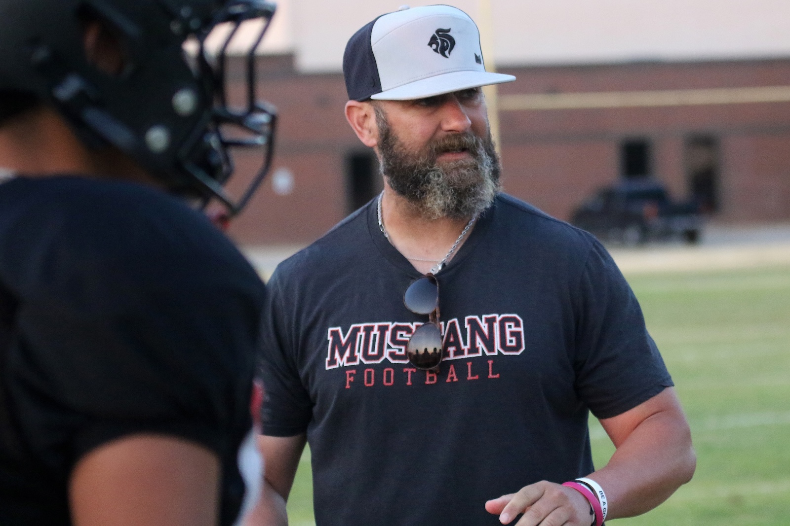 Bearded man in a Mustang Football shirt and white/navy cap stands on a football field, sunglasses hung from his shirt.