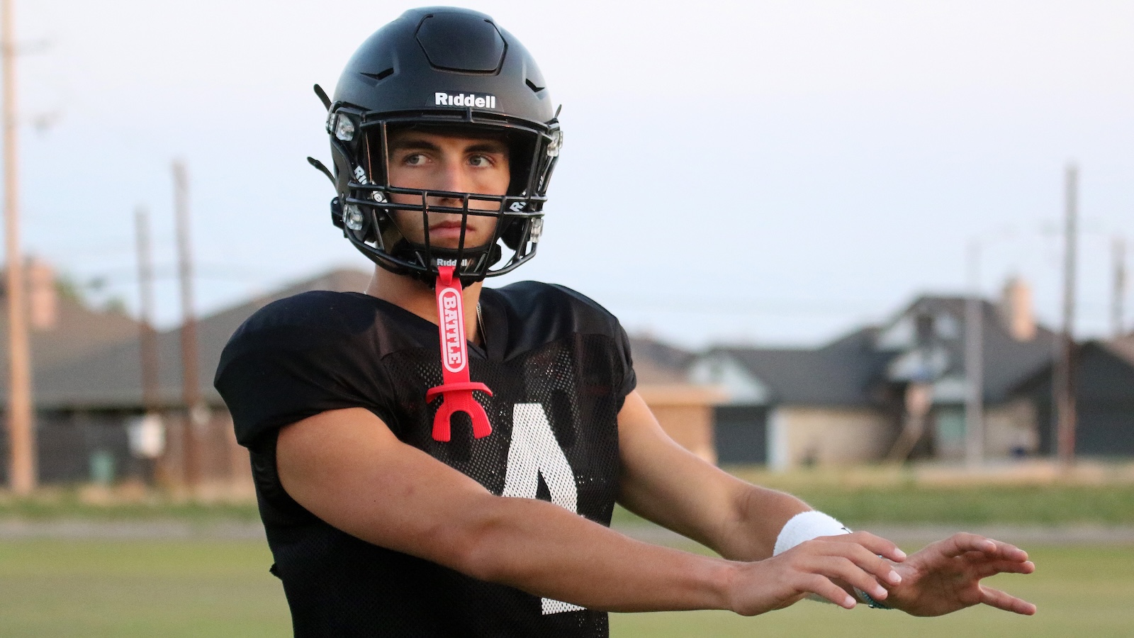 Youth football player in a black jersey and Riddell helmet on a grassy field, red mouthguard hanging from facemask, suburban homes in the background.