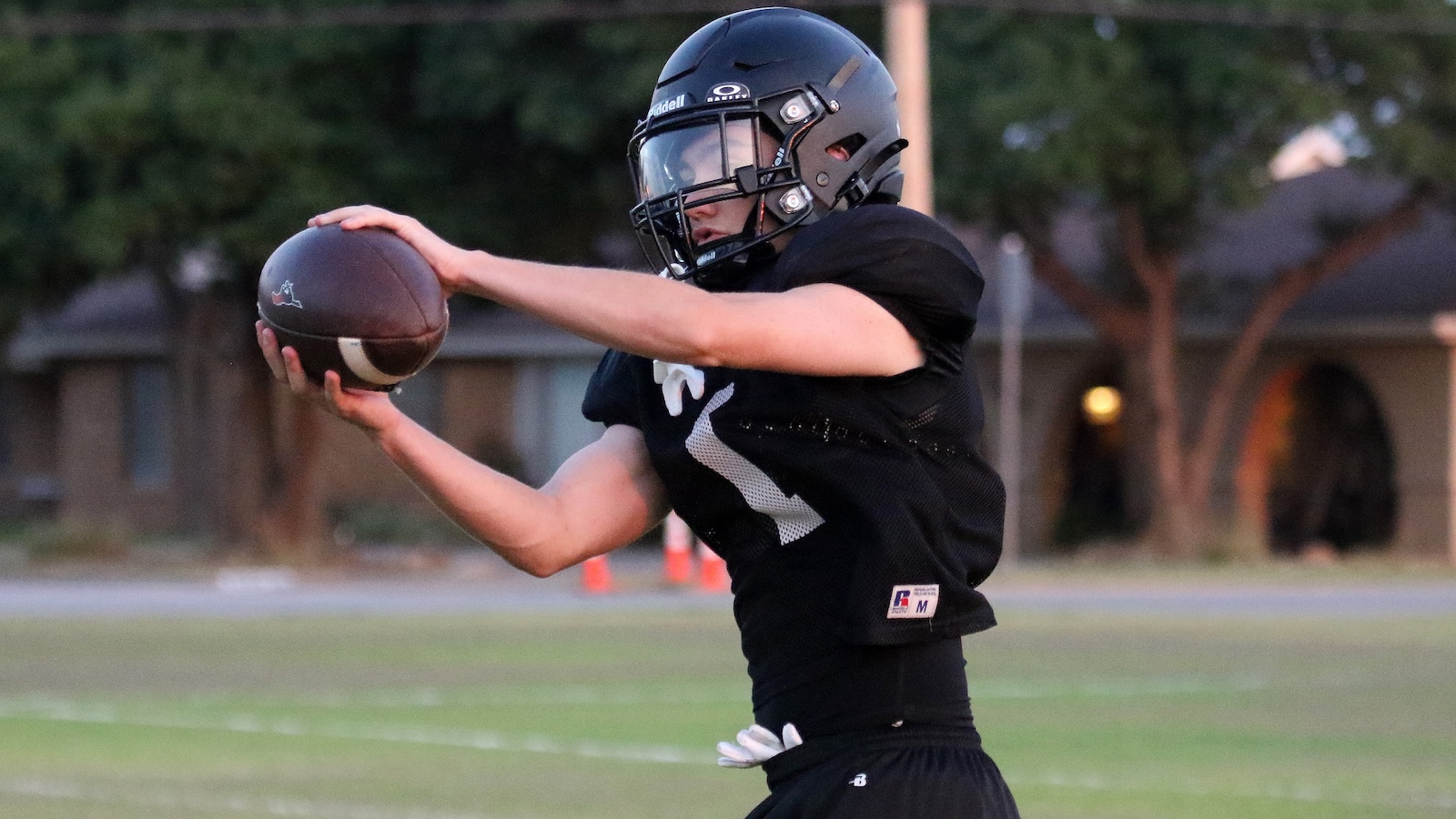 Football player in a black jersey and helmet reaching to catch a football on a grassy field.
