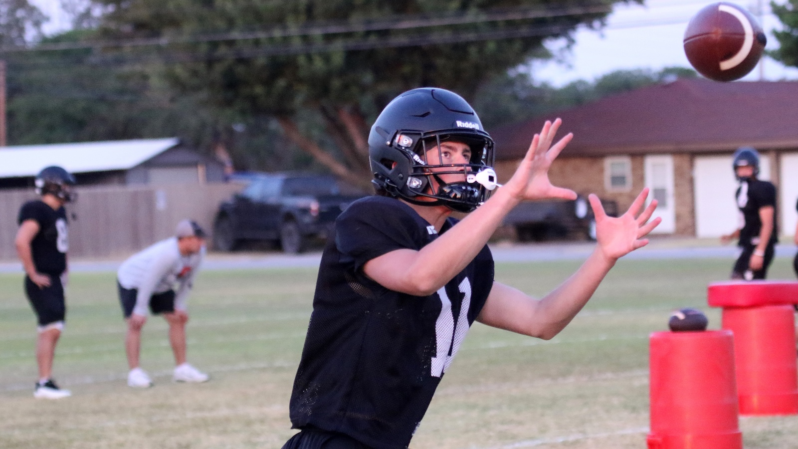 Youth football player in black jersey and helmet reaches to catch a spiraling football during practice on a grassy field.