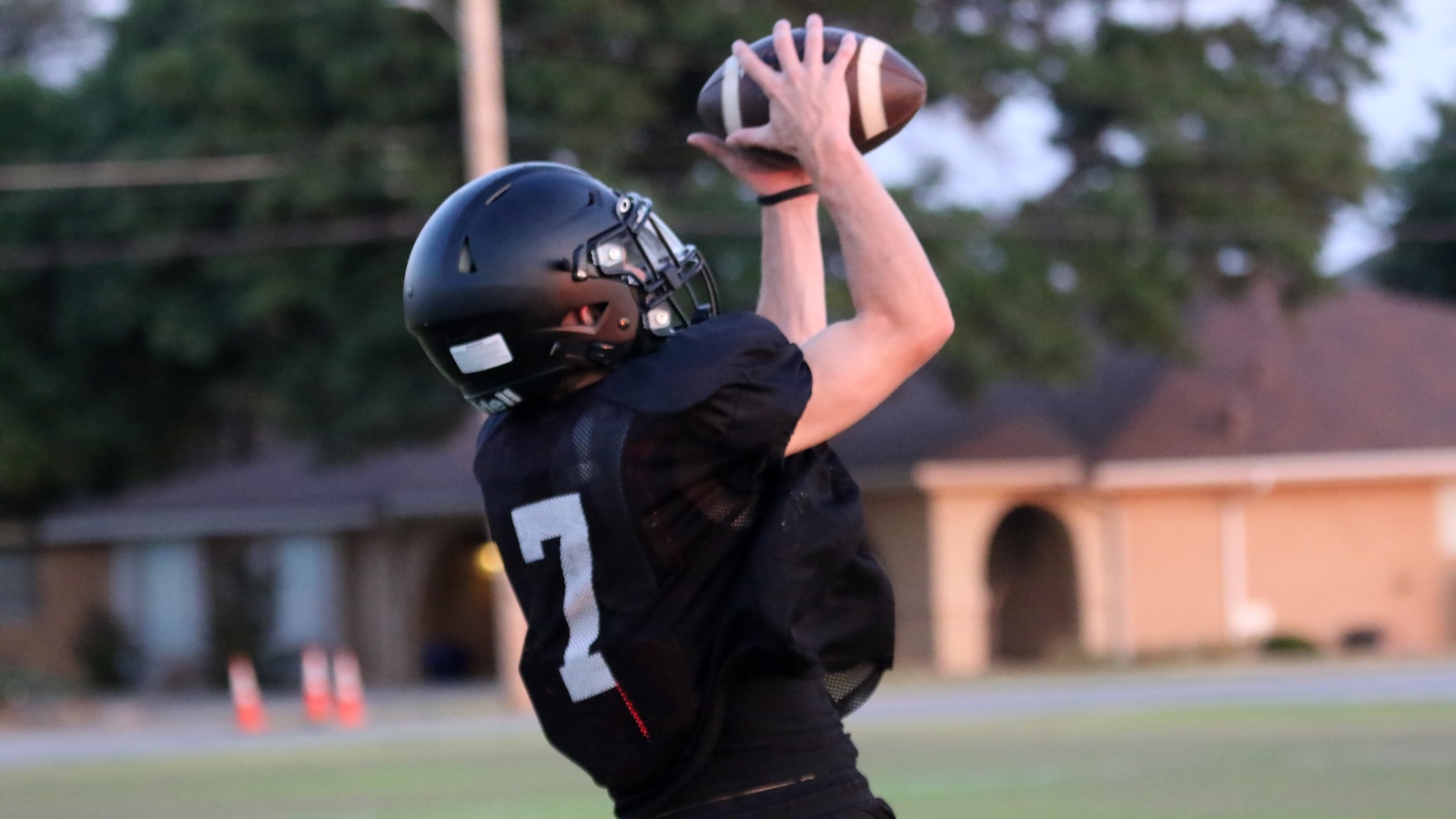 Football player in a black jersey #7 reaches up to catch a football in midair, wearing a black helmet on a sports field.