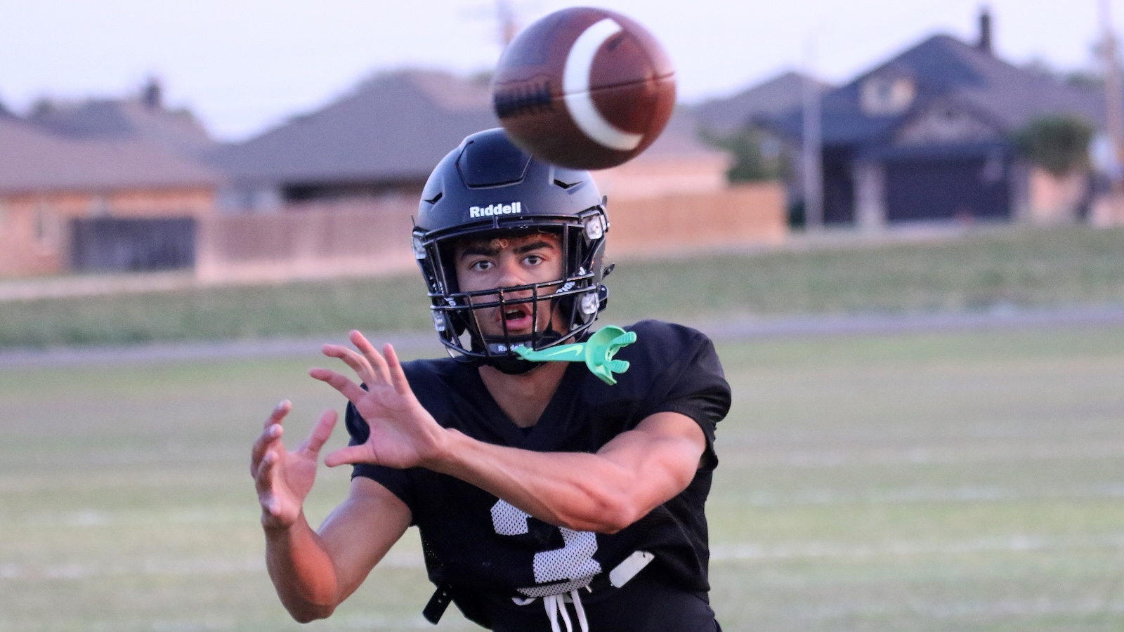 Male football player in a black jersey and helmet extends hands to catch a brown football mid-air on a grassy field.