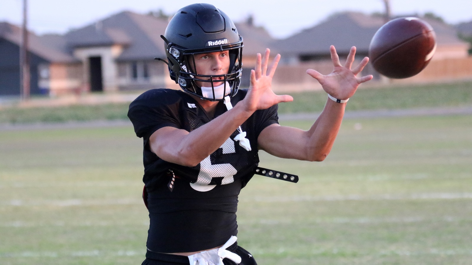American football player in a black uniform and helmet reaches to catch a ball on a practice field