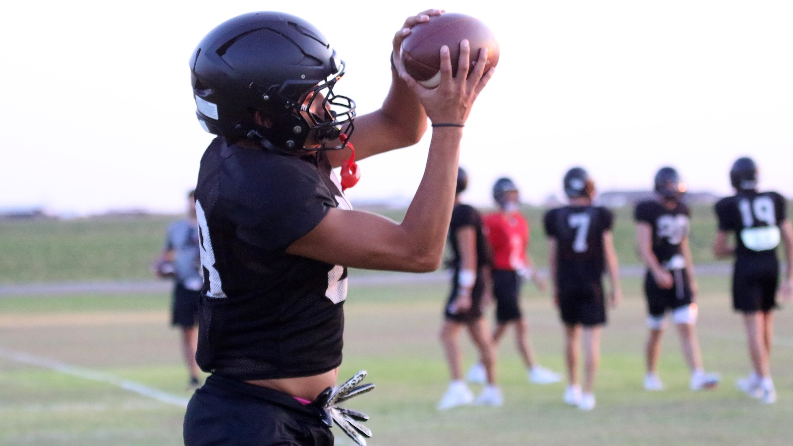 Football player in a black uniform catches a football during practice on a grassy field, teammates blurred in the background.