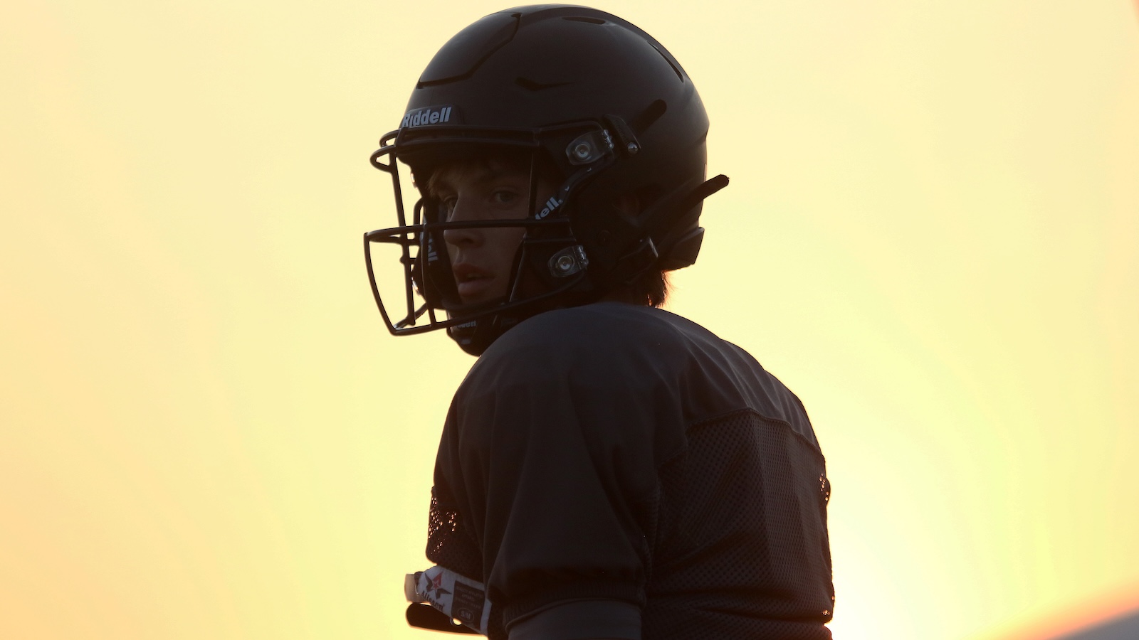American football player in a black helmet and dark jersey looking over his shoulder at sunset.
