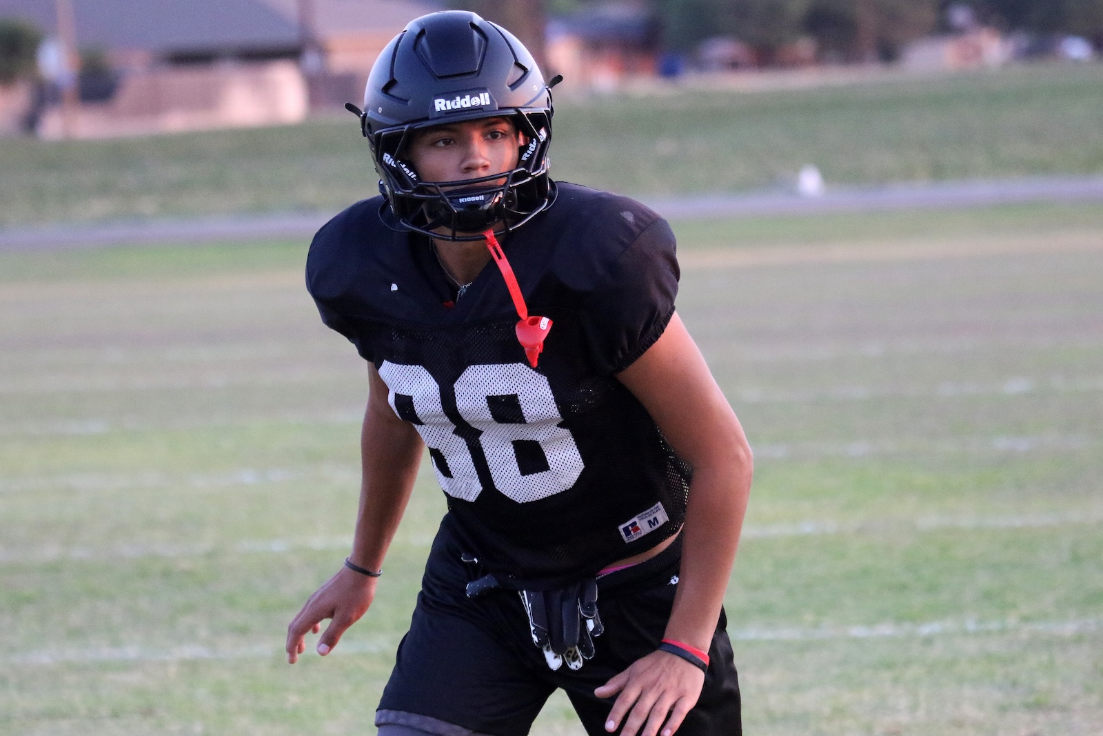 Youth football player in a black jersey with number 88, wearing a helmet, ready on a grassy field with a red whistle hanging from the neck.