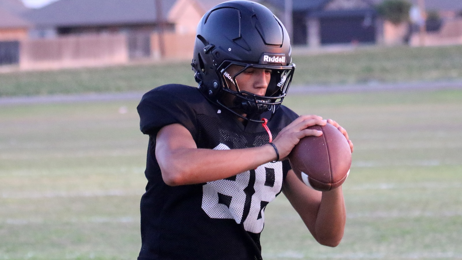 Young football player in a black jersey and helmet grips a football, preparing to throw on a grassy field.