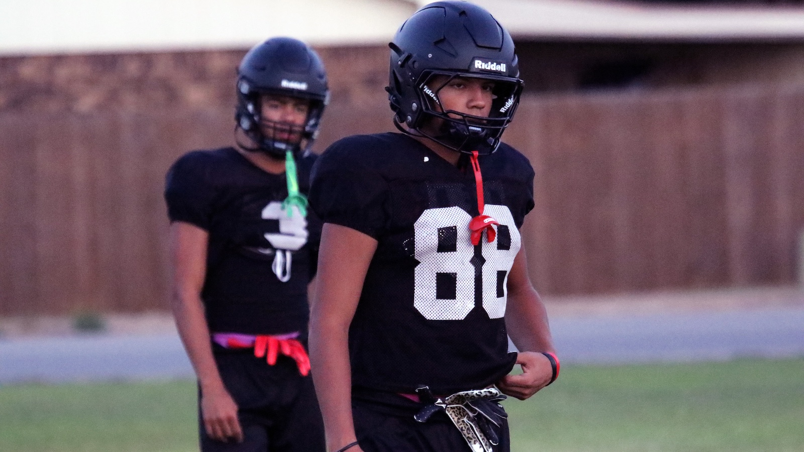 Two football players in black uniforms and Riddell helmets stand on a practice field, one in front wearing a red lanyard.