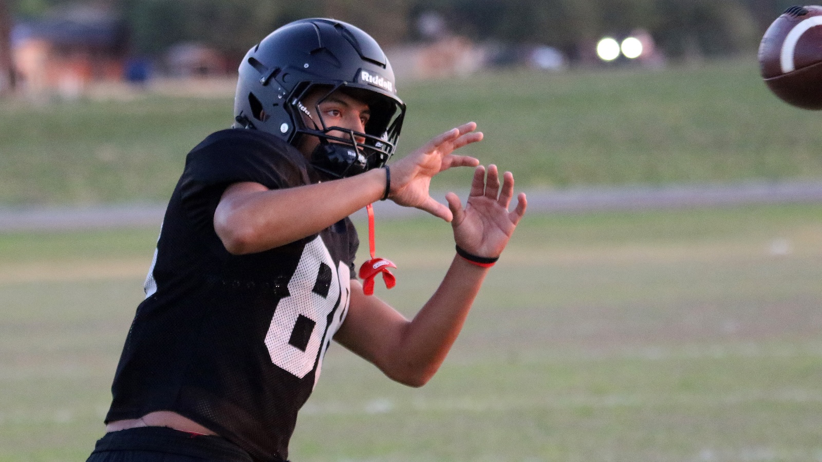Youth football player in a black jersey and helmet reaches out to catch an incoming ball during a game on a grassy field.