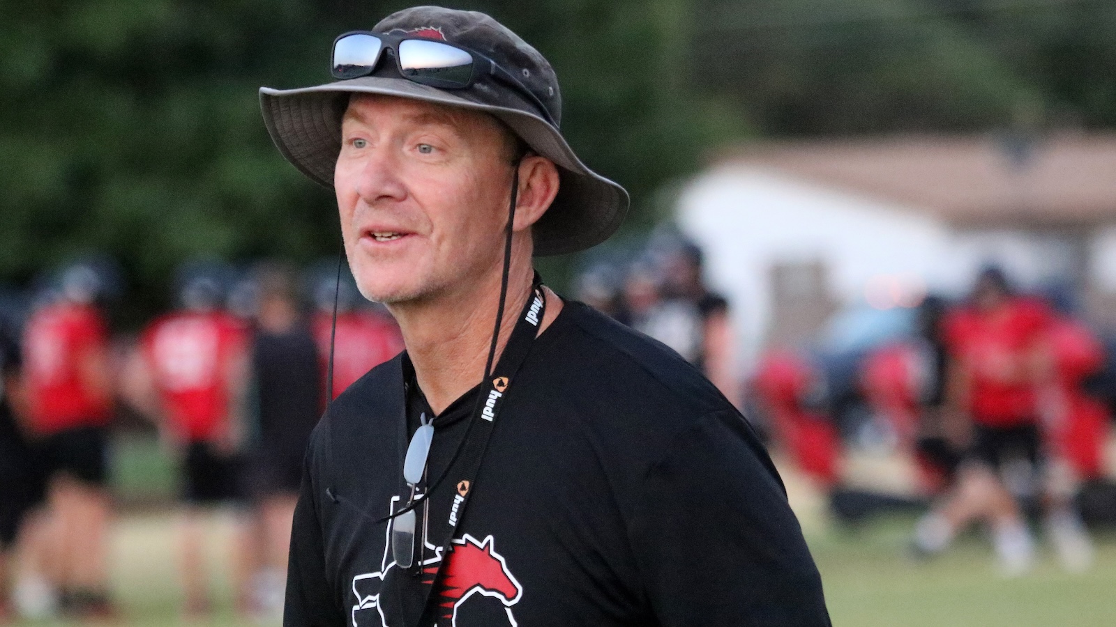 Man wearing a bucket hat and lanyard at an outdoor sports event, with blurred people in red shirts in the background.