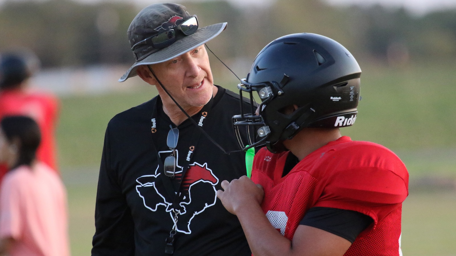 Football coach wearing a hat and sunglasses speaks with a player in a red jersey and helmet during practice on a field.
