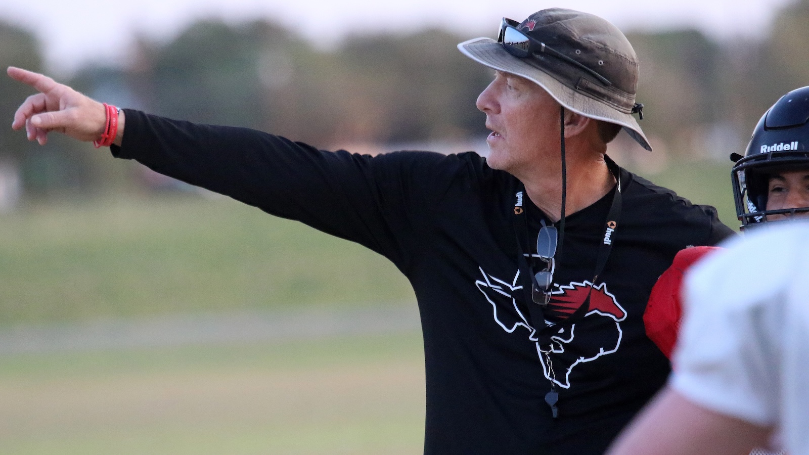 Coach on a field, wearing a bucket hat and black shirt, points to the left as players watch nearby.