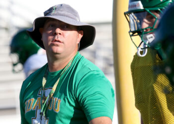Idalou - HC Clay White 3 | Press Pass Sports Man in a green shirt wearing a wide-brimmed hat and lanyard, standing by a football field with a helmeted teammate nearby.