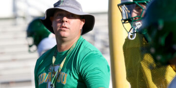 Man in a green shirt wearing a wide-brimmed hat and lanyard, standing by a football field with a helmeted teammate nearby.