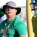 Man in a green shirt wearing a wide-brimmed hat and lanyard, standing by a football field with a helmeted teammate nearby.