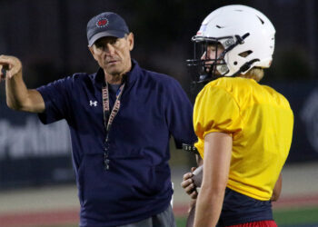 Liberty - HC Joe Sexton 1 | Press Pass Sports Coach in a navy shirt and cap gestures as he talks to a football player in a yellow jersey and white helmet on a dimly lit field at night.