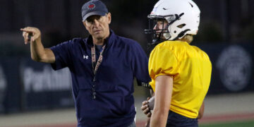 Coach in a navy shirt and cap gestures as he talks to a football player in a yellow jersey and white helmet on a dimly lit field at night.