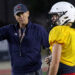 Liberty - HC Joe Sexton 1 | Press Pass Sports Coach in a navy shirt and cap gestures as he talks to a football player in a yellow jersey and white helmet on a dimly lit field at night.