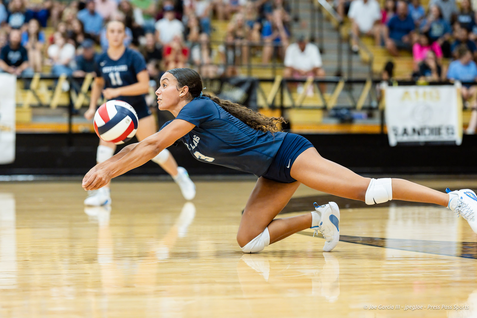 west-plains-vs-amarillo-high-2026-jpegJoe-pps-2 | Press Pass Sports Female volleyball player in a navy uniform lunges on the court to reach a fast-moving ball, with spectators in the stands behind her.
