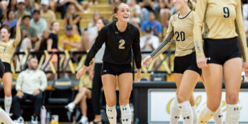 Volleyball players on the court celebrate; a player in black with number 2 smiles as teammates in beige jerseys (numbers 20 and 9) stand nearby, cheering crowd in the stands behind.