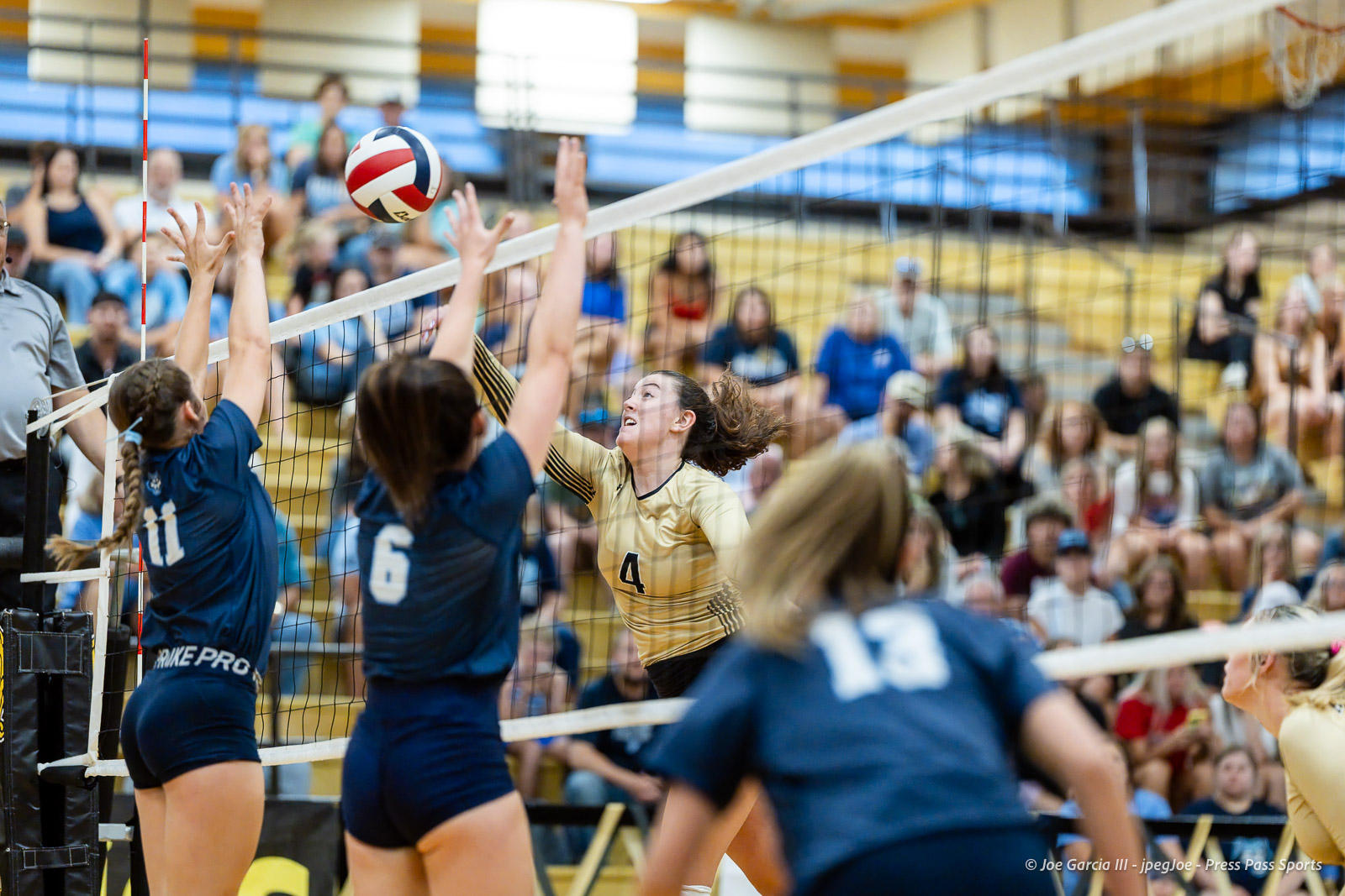 west-plains-vs-amarillo-high-2026-jpegJoe-pps-14 | Press Pass Sports Gold-team player wearing #4 spikes the ball over the net as blue-team blockers jump to defend; crowded gym audience in bleachers watches closely.