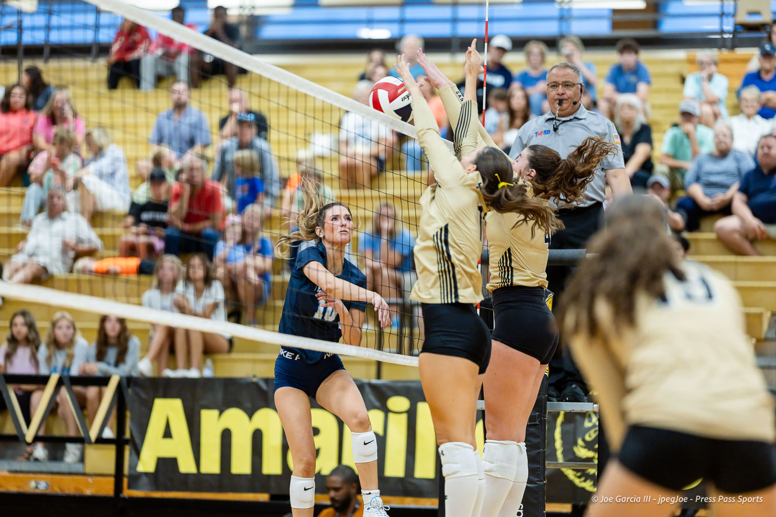 west-plains-vs-amarillo-high-2026-jpegJoe-pps-16 | Press Pass Sports Female volleyball players jump to block the ball at the net as a teammate in blue watches from below in a gym filled with spectators on bleachers.