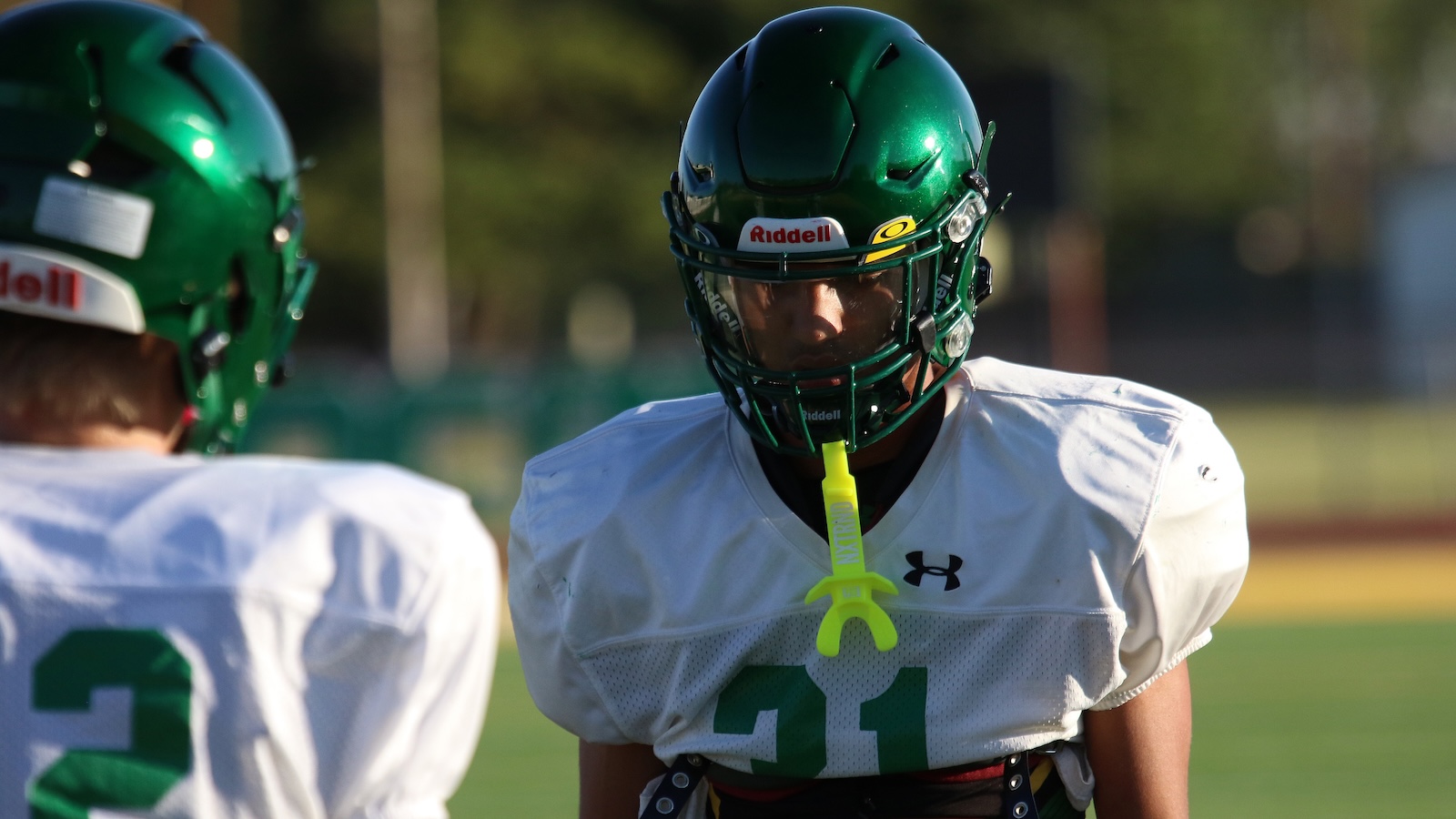 Two football players in green helmets and white jerseys on a field; foreground player wears a bright neon mouthguard and number 31, facing the camera (practice scene).