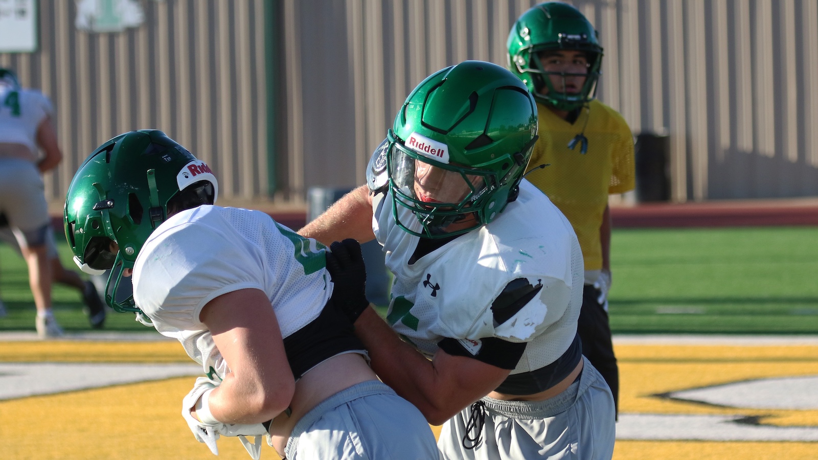 Two football players in green helmets tackle each other on a yellow turf field, white jerseys and one in a yellow background watching.