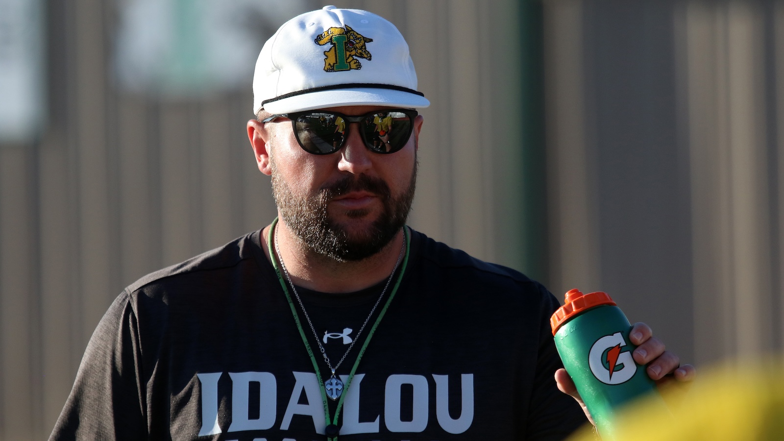 Man with a white cap and sunglasses, bearded, holding a green Gatorade bottle outdoors.