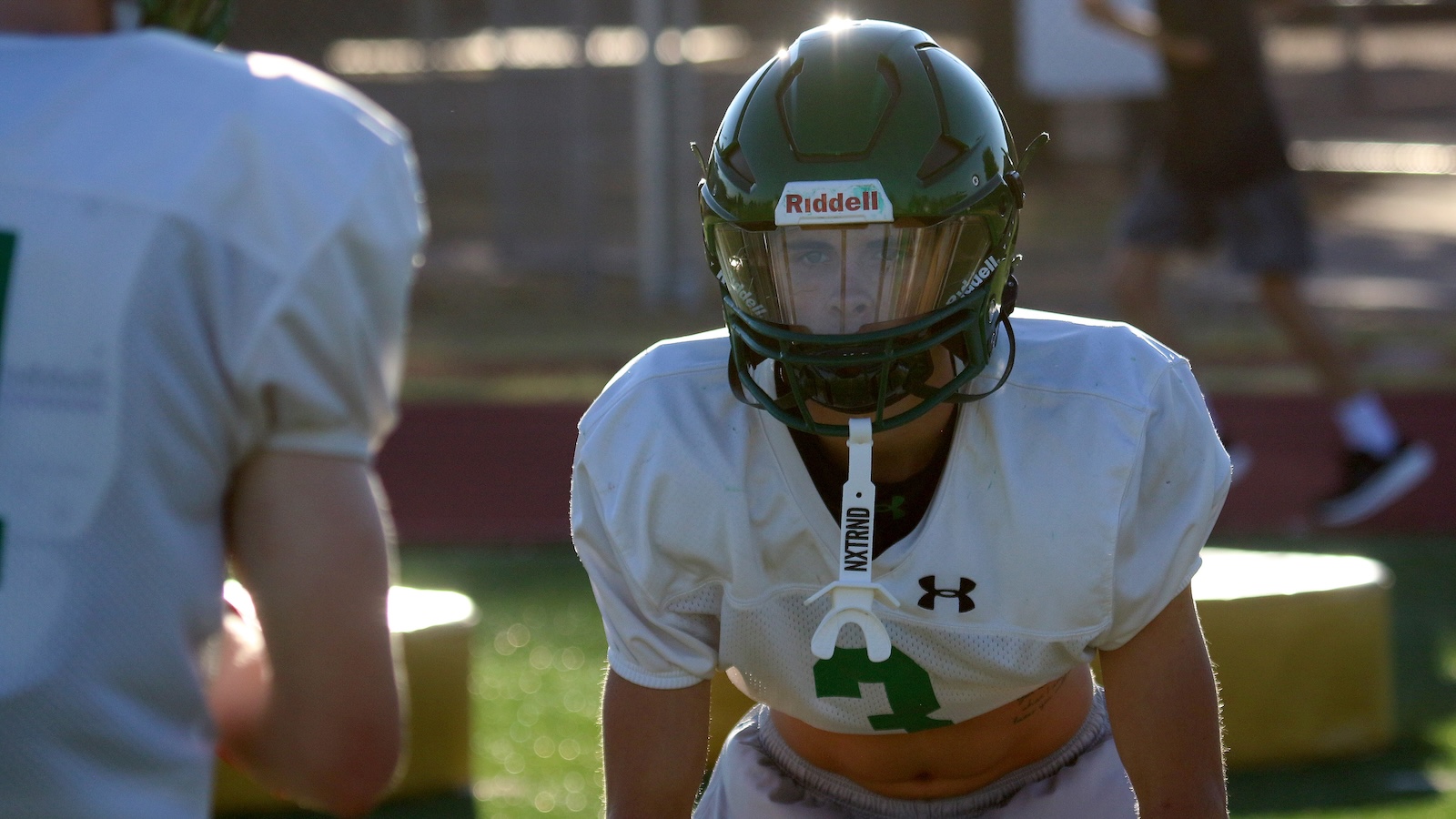 Football player in a white jersey and green helmet crouches in a drill on a sunny practice field, facing the camera.