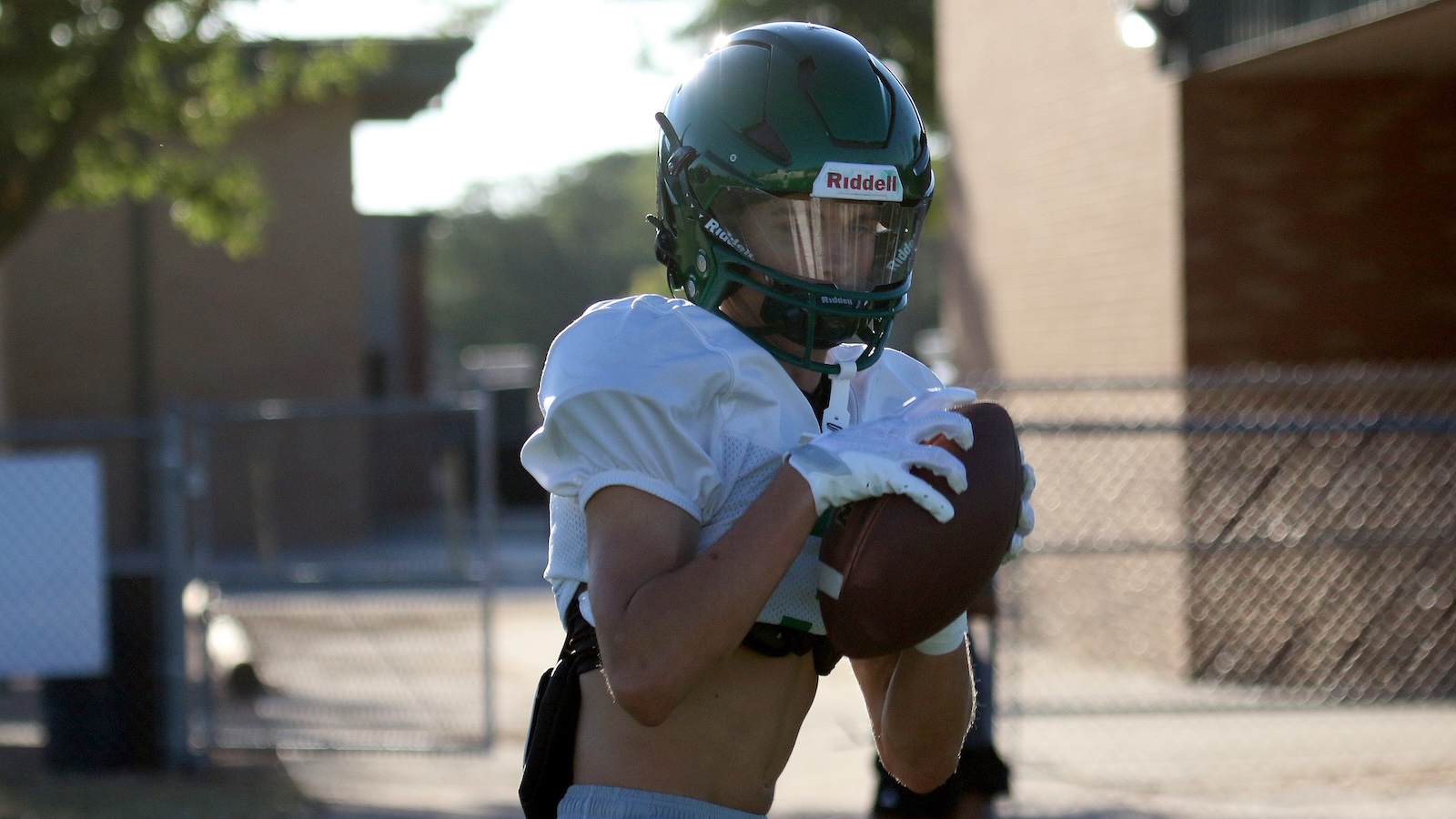 Football player in a green helmet and white jersey cradling a brown ball, preparing to run outdoors near a chain-link fence.