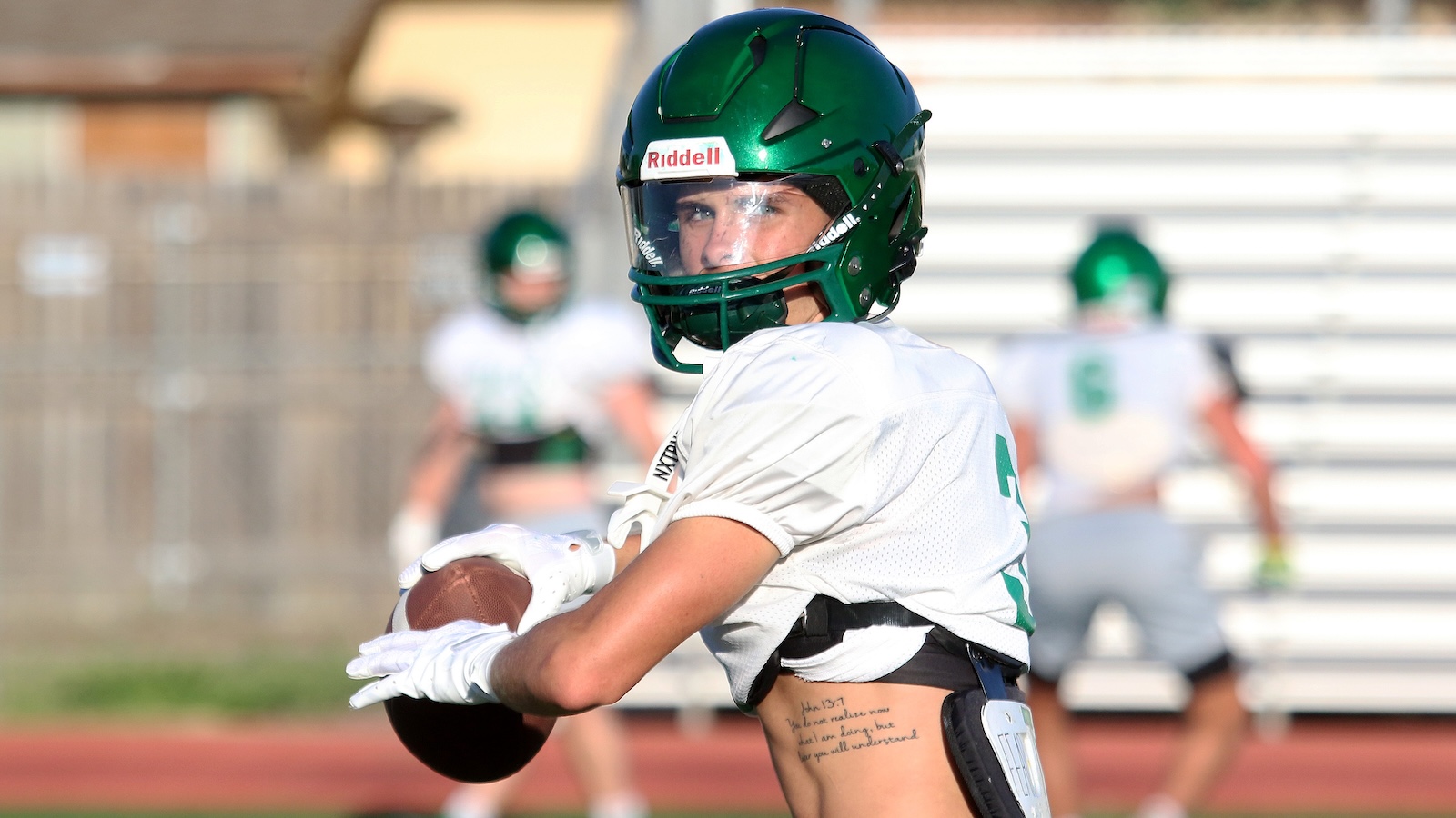 Football player in white jersey and green helmet catching a football on a sunny practice field.