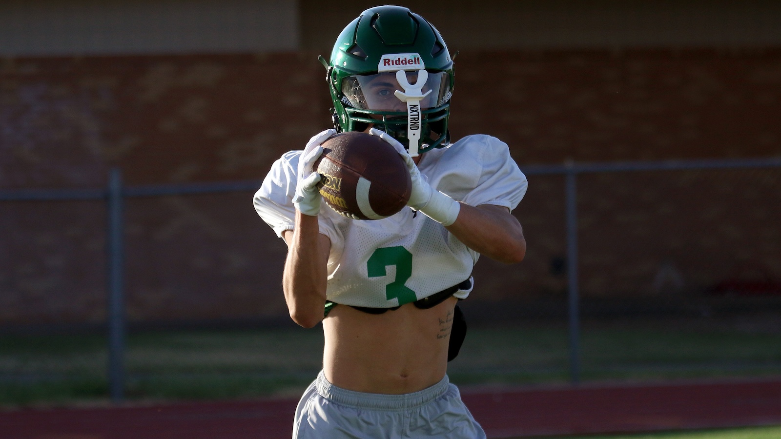 Football player in a white jersey with number 3, green helmet, catching a football mid-play on a field graphic backdrop.