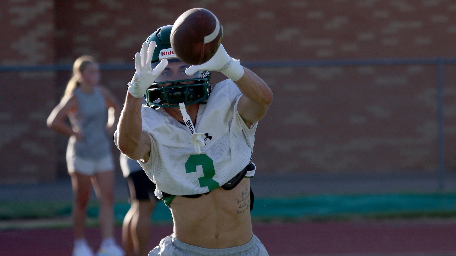 Football player in a white jersey with green number 3 reaches to catch a football, with two teammates blurred in the background.